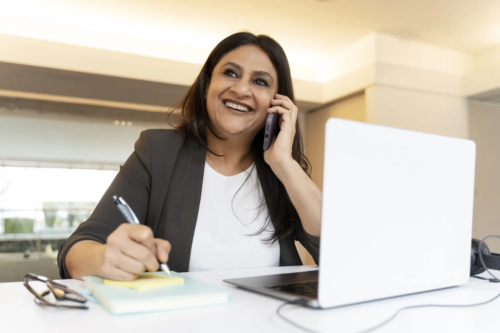 Asian business woman talking on a mobile phone while writing notes at a desk with a laptop.