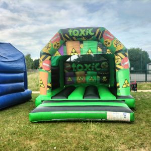 Inflatable bounce house named 'Toxic Castle' featuring colorful hazard symbols and a green base.