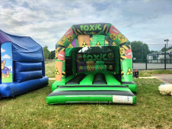 Inflatable bounce house named 'Toxic Castle' featuring colorful hazard symbols and a green base.